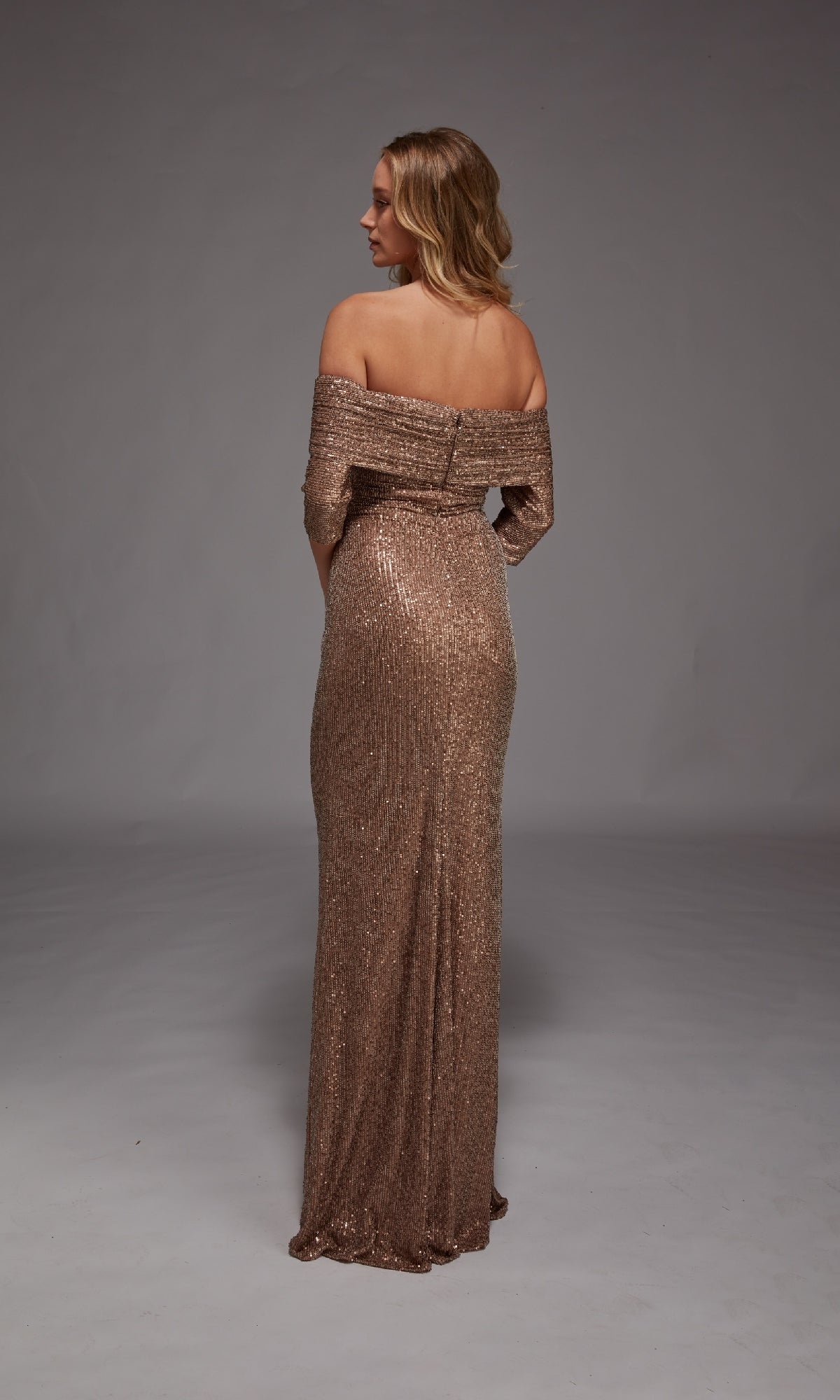 A woman stands facing away, wearing the Alyce 27787 Long Formal Dress—a floor-length, off-the-shoulder mother-of-the-bride gown in light gold sequins—against a plain gray background.